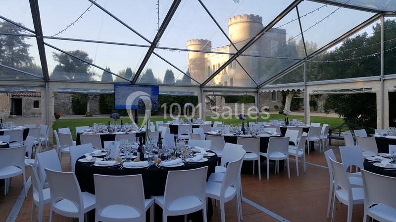 Salle de réception sous une tente transparente avec tables dressées, vue sur un château en arrière-plan.