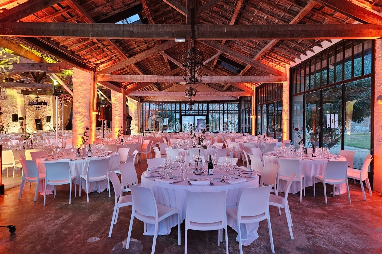 Salle de réception avec tables rondes dressées, chaises blanches et éclairage chaleureux sous une charpente en bois.