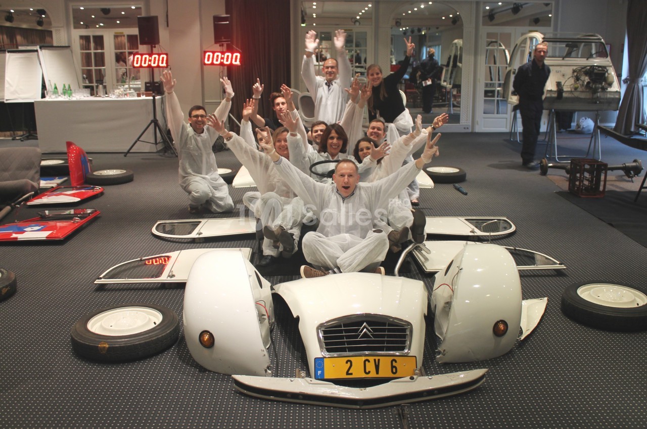 Des personnes en combinaison blanche posent joyeusement autour d'une voiture Citroën 2CV démontée dans une salle d…