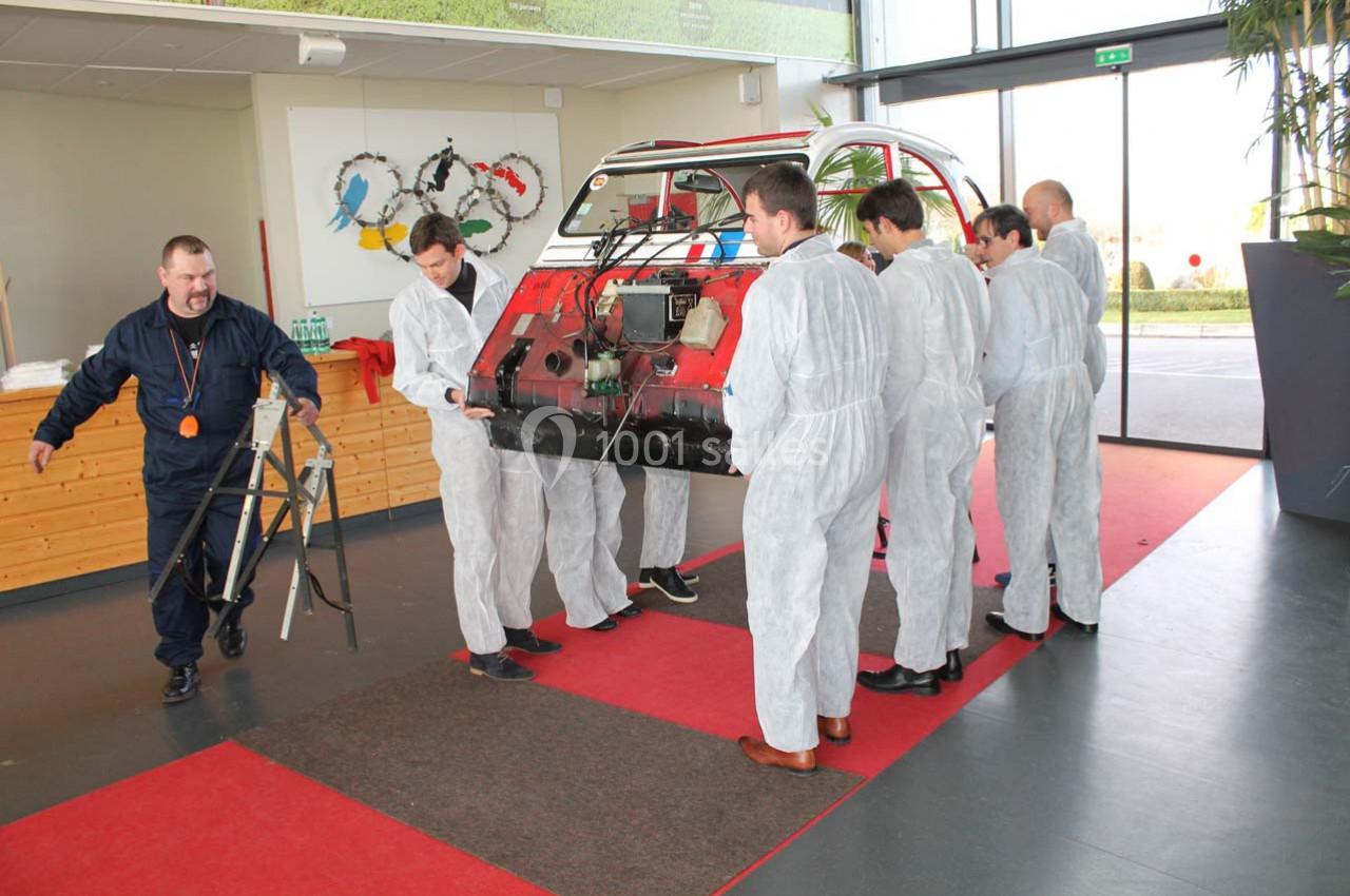 Des personnes en combinaison blanche manipulent une grande pièce de carrosserie rouge dans un espace intérieur lumineux.