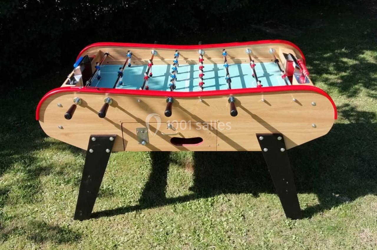 Baby-foot en bois avec surface de jeu bleue et joueurs rouges et bleus, posé sur une pelouse verte.