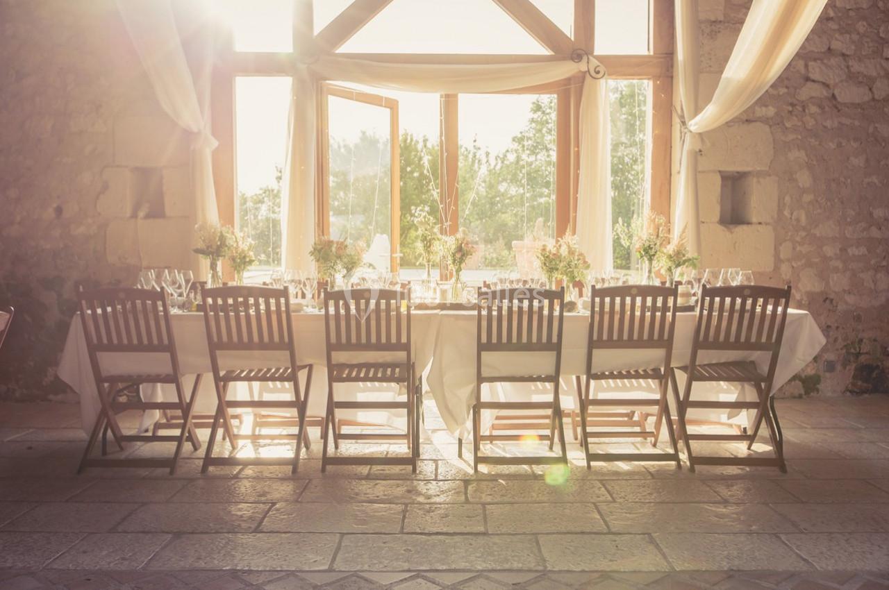Grande table dressée avec des chaises en bois, décorée de fleurs, dans une salle lumineuse avec de grandes fenêtres.