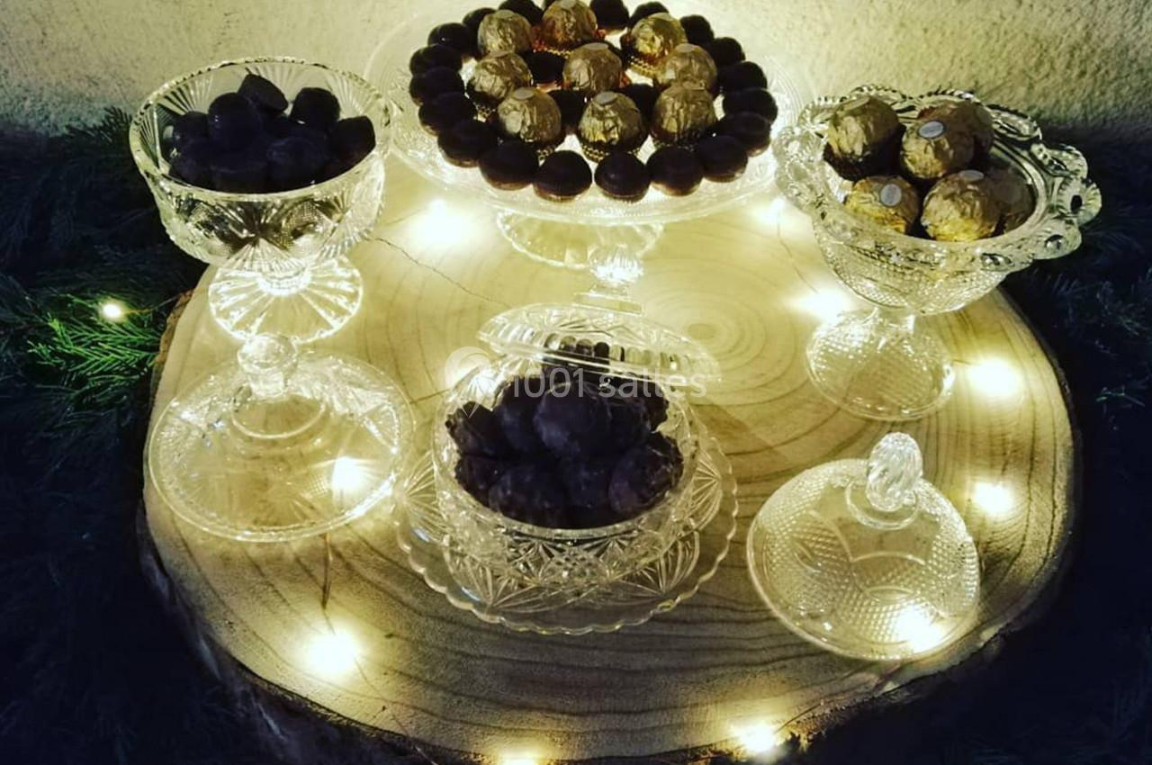 Assortiment de chocolats présentés dans des coupes en verre sur une tranche de bois éclairée par des guirlandes lumineuses.