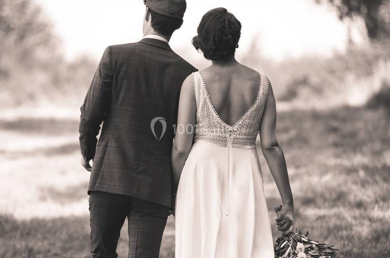 Un couple de dos, vêtu élégamment, marche dans un cadre naturel, la femme tenant un bouquet de fleurs.