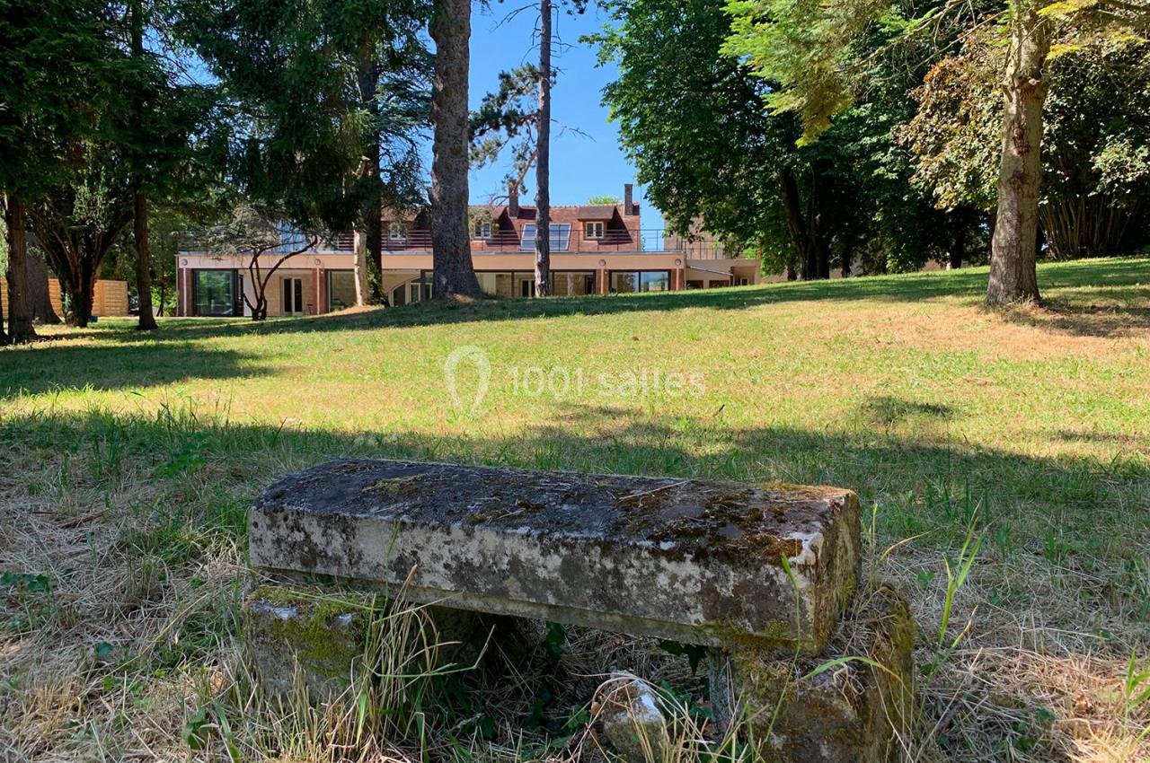 Location salle Monceaux (Oise) - Domaine de Monceaux #10 Banc en pierre ancienne dans un parc verdoyant, avec une maison moderne visible en arrière-plan.