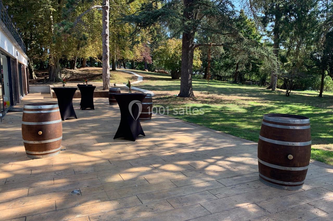 Location salle Monceaux (Oise) - Domaine de Monceaux #15 Terrasse en bois avec des tonneaux et des tables hautes noires, entourée d'arbres dans un cadre verdoyant.