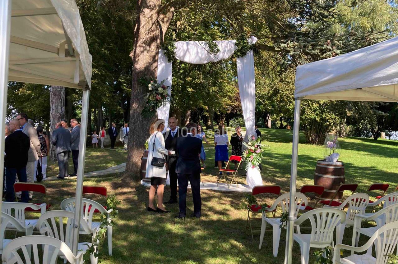 Location salle Monceaux (Oise) - Domaine de Monceaux #16 Cérémonie en plein air sous des arbres, avec des chaises blanches disposées et un arche décoré de tissu blanc.
