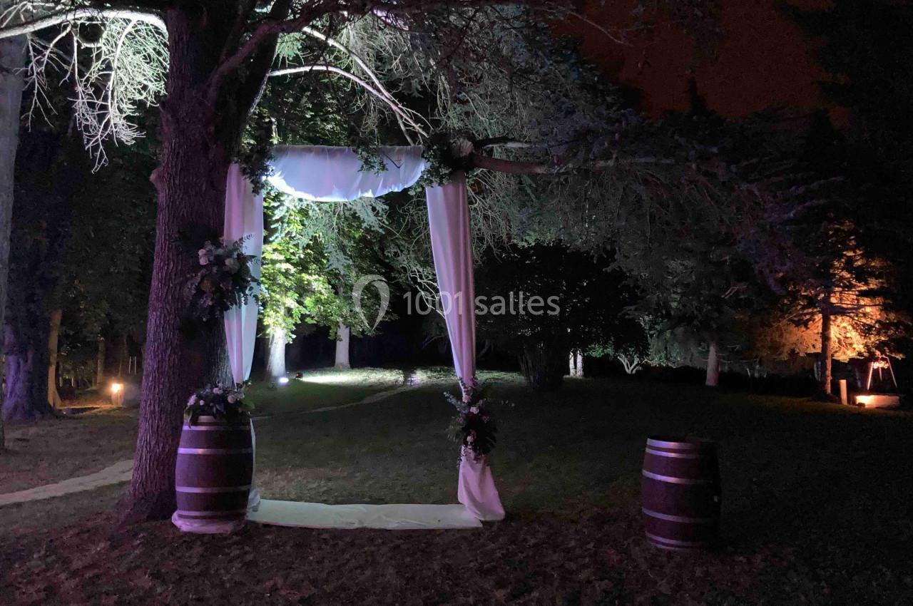 Location salle Monceaux (Oise) - Domaine de Monceaux #18 Arche de mariage décorée de voilages blancs et de fleurs, installée en extérieur sous des arbres éclairés la nuit.