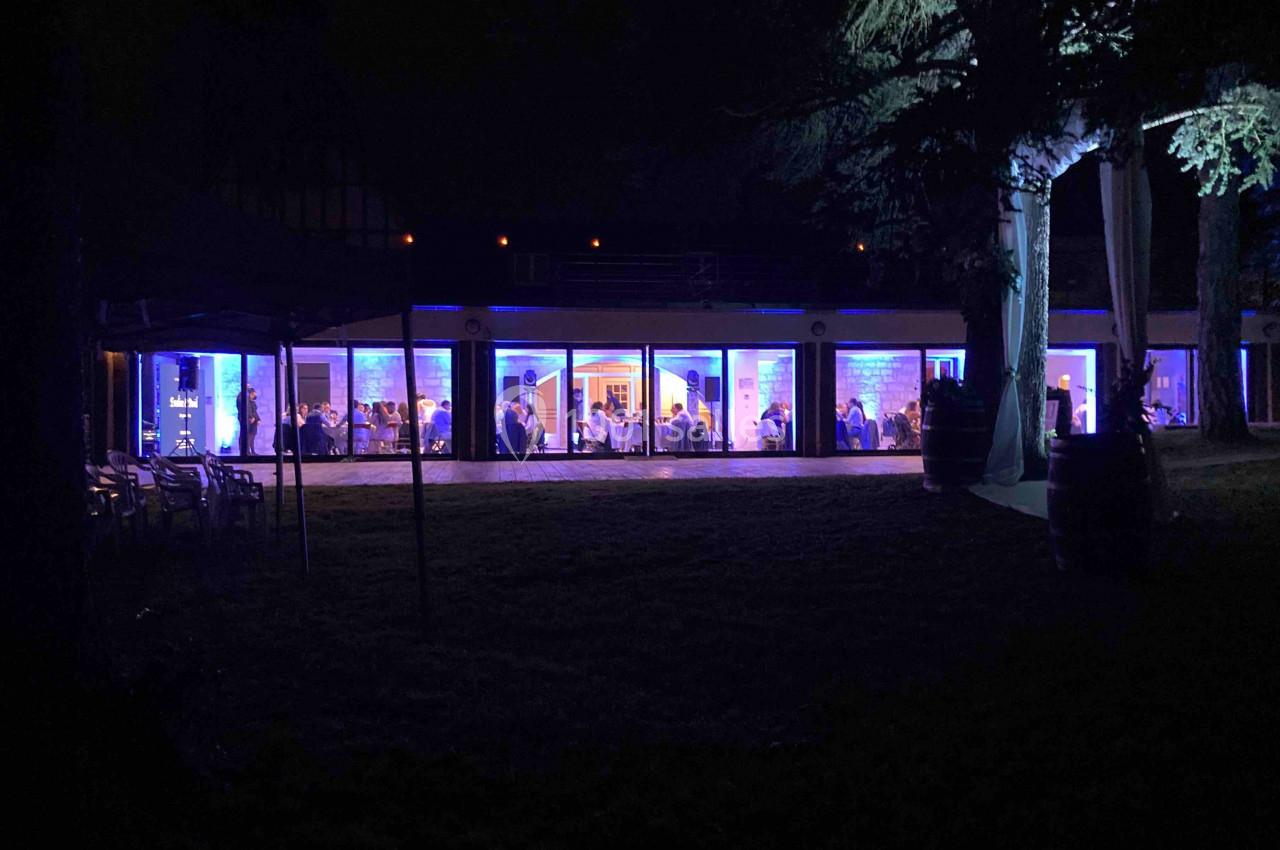 Location salle Monceaux (Oise) - Domaine de Monceaux #20 Salle vitrée éclairée en bleu la nuit, avec des personnes visibles à l'intérieur et des arbres en premier plan.