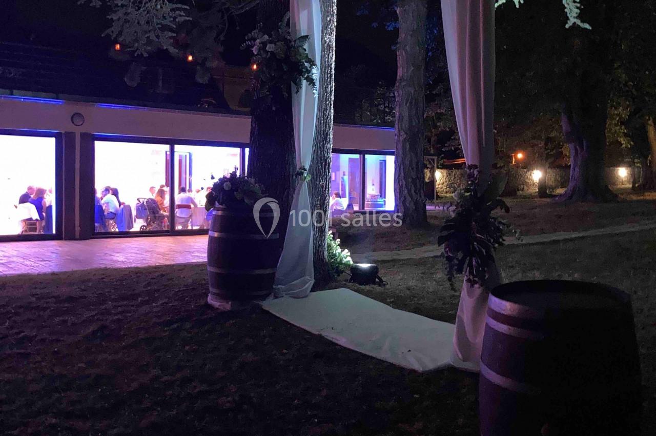 Location salle Monceaux (Oise) - Domaine de Monceaux #21 Décoration de mariage en extérieur avec arche lumineuse sous un arbre, barils en bois et salle éclairée en arrière-plan.