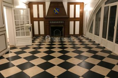 Salle avec un sol en damier noir et beige, cheminée en bois, murs blancs et grandes fenêtres cintrées.