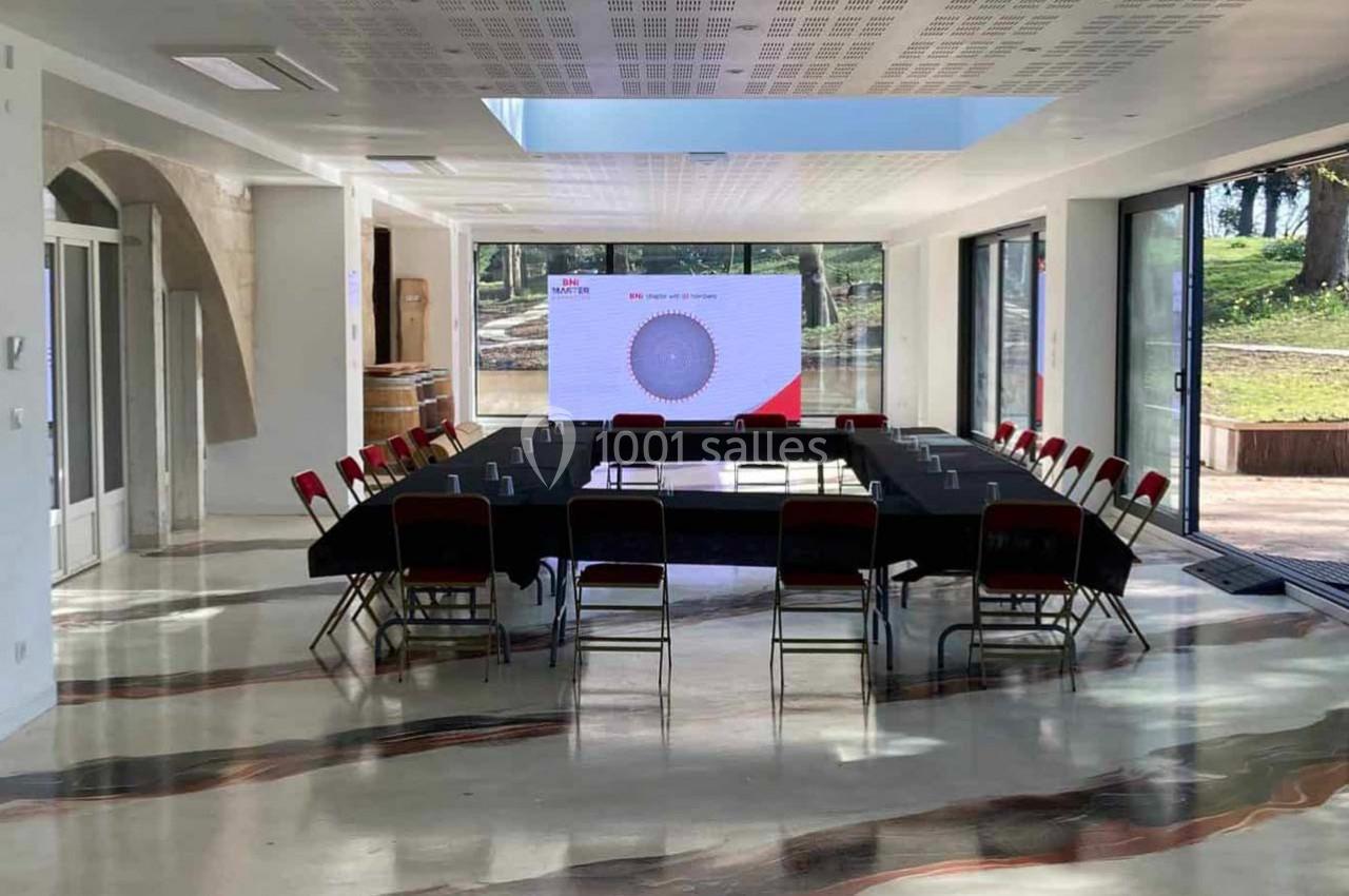 Salle de réunion lumineuse avec une grande table en U, des chaises rouges et un écran de présentation allumé.
