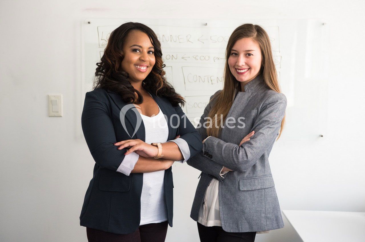 Deux femmes souriantes debout devant un tableau blanc, bras croisés, dans un environnement professionnel.