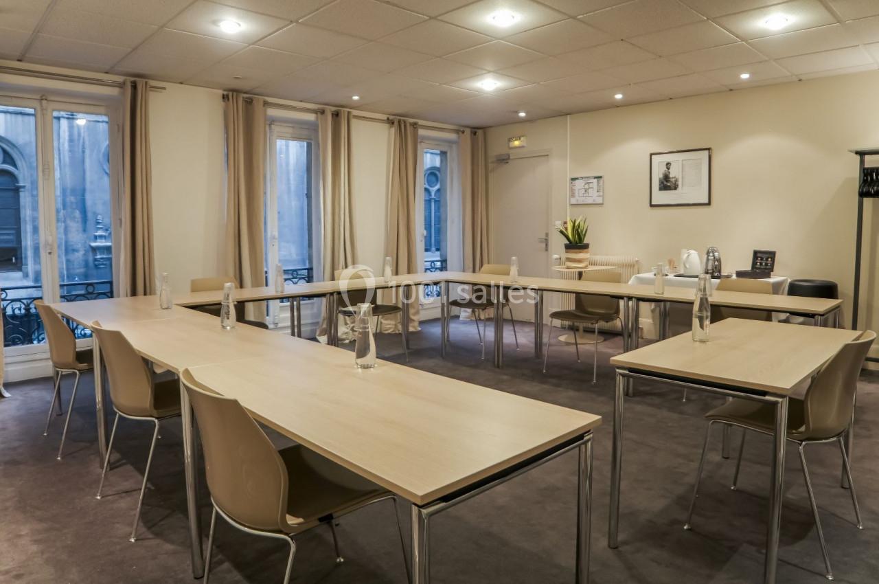 Salle de réunion lumineuse avec tables en bois disposées en U, chaises beige et fenêtres donnant sur l'extérieur.