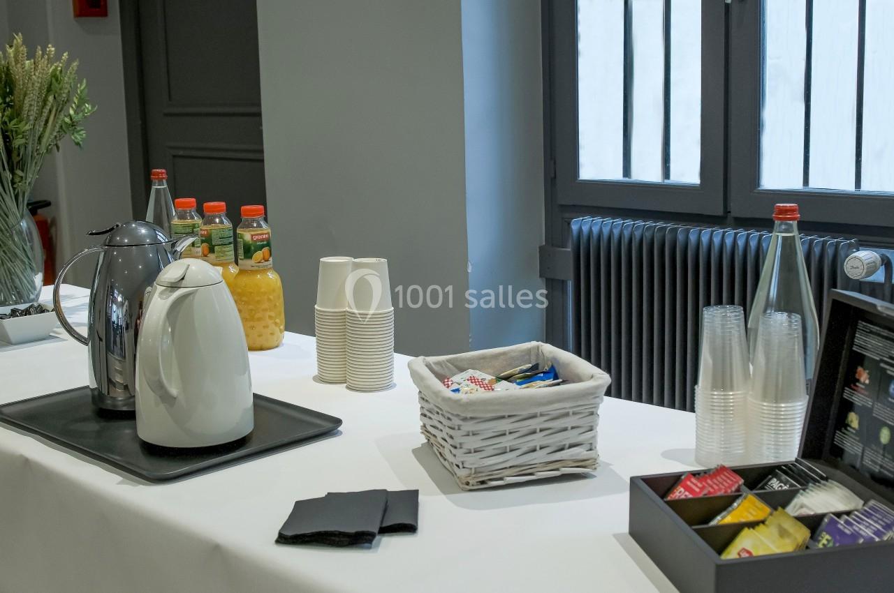 Table avec boissons, gobelets, thé, café, jus d'orange et accessoires, disposée près d'une fenêtre.