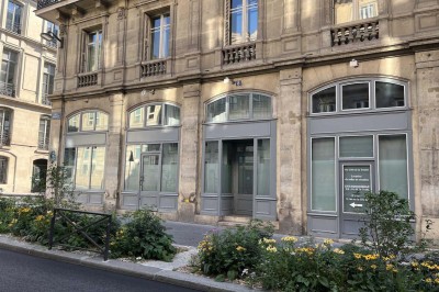 Façade d'un immeuble en pierre avec des fenêtres vitrées et un parterre de fleurs en bordure de trottoir.