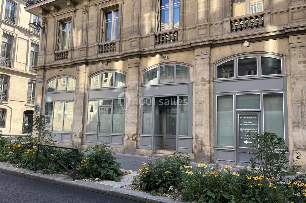 Façade d'un immeuble en pierre avec des fenêtres vitrées et un parterre de fleurs en bordure de trottoir.