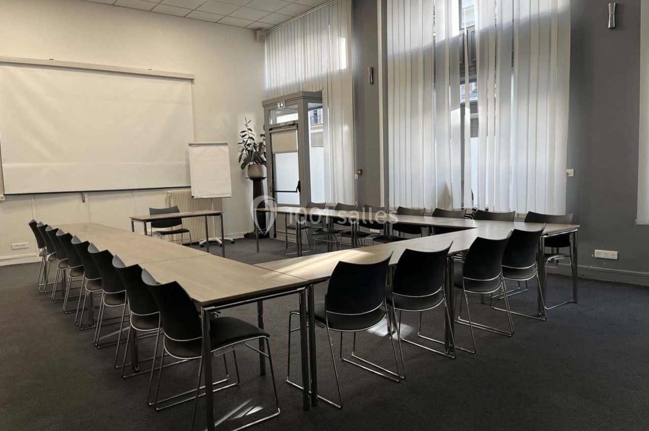 Salle de réunion lumineuse avec tables disposées en U, chaises noires et écran de projection.
