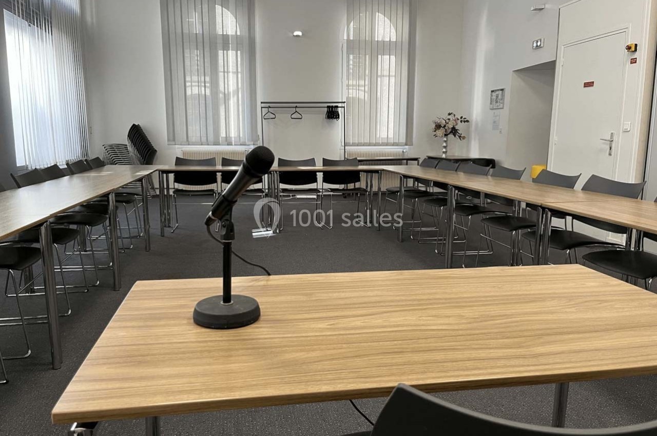 Salle de réunion lumineuse avec tables disposées en U, un micro sur une table et des chaises alignées.