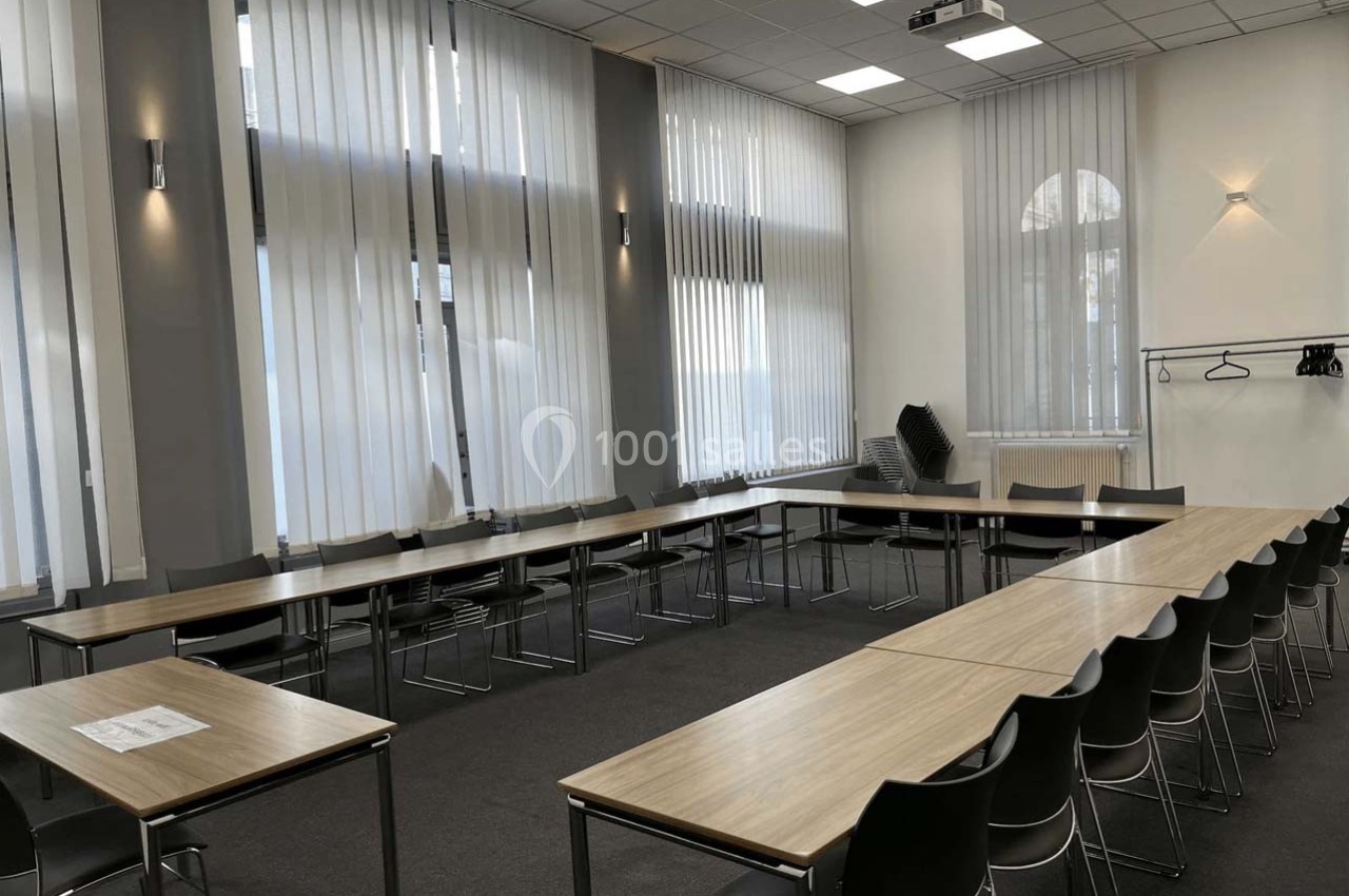 Salle de réunion lumineuse avec des tables en U, des chaises noires, des rideaux translucides et un portemanteau.