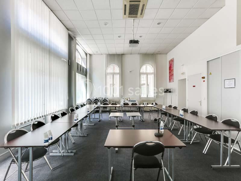 Salle de réunion lumineuse avec tables en U, chaises noires, projecteur au plafond et grandes fenêtres avec stores.