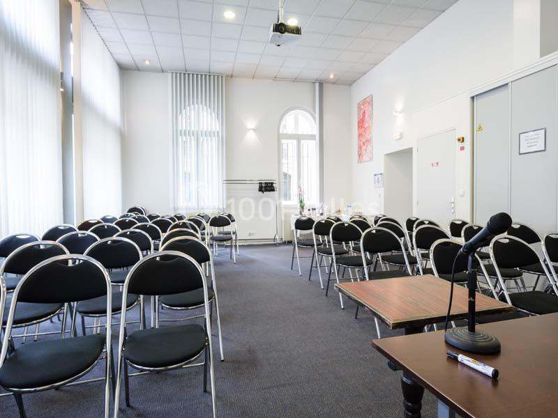 Salle de conférence lumineuse avec des chaises noires alignées, un micro sur une table et un projecteur au plafond.