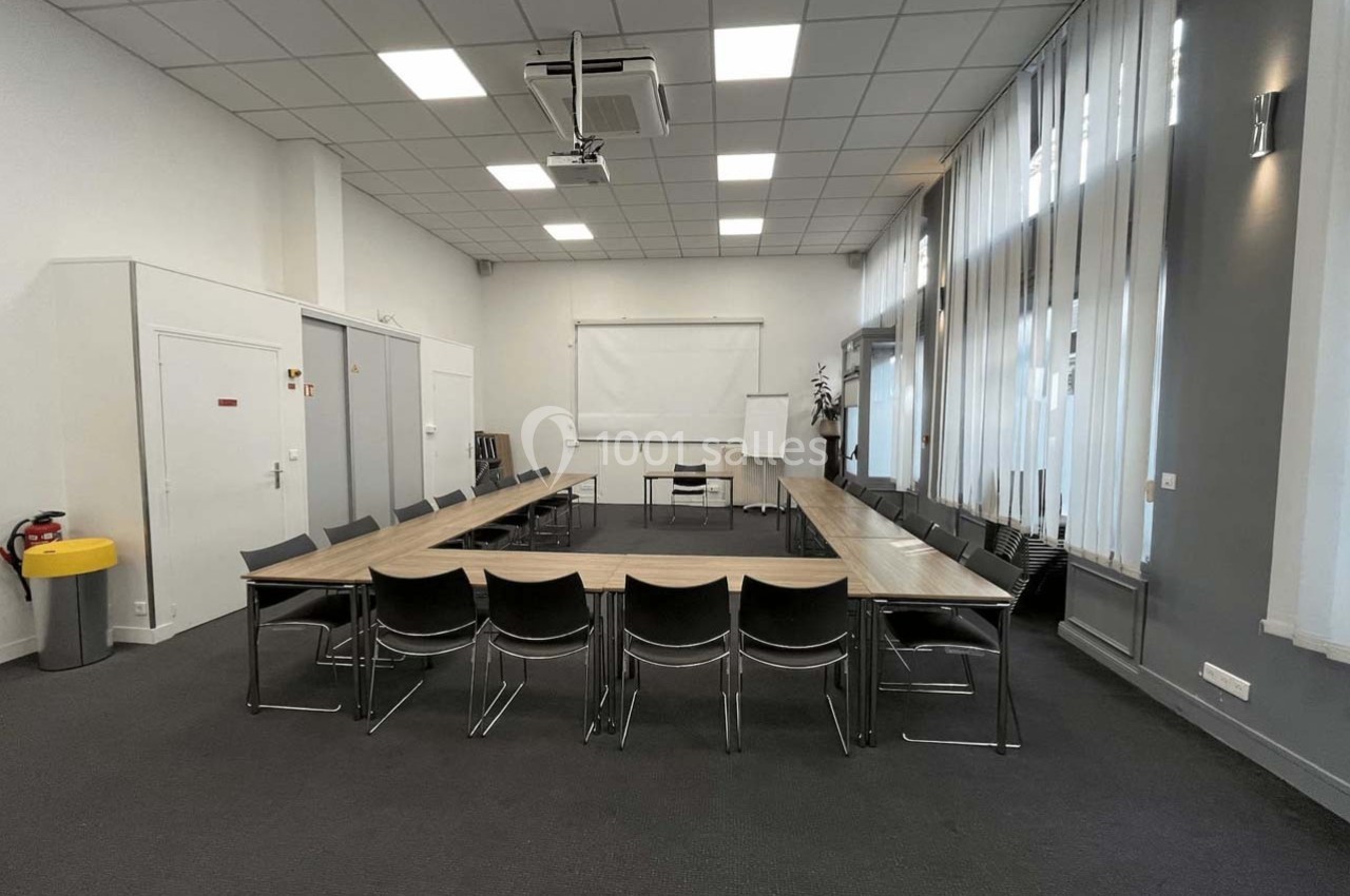 Salle de réunion avec tables disposées en U, chaises noires, tableau blanc et grandes fenêtres avec rideaux.