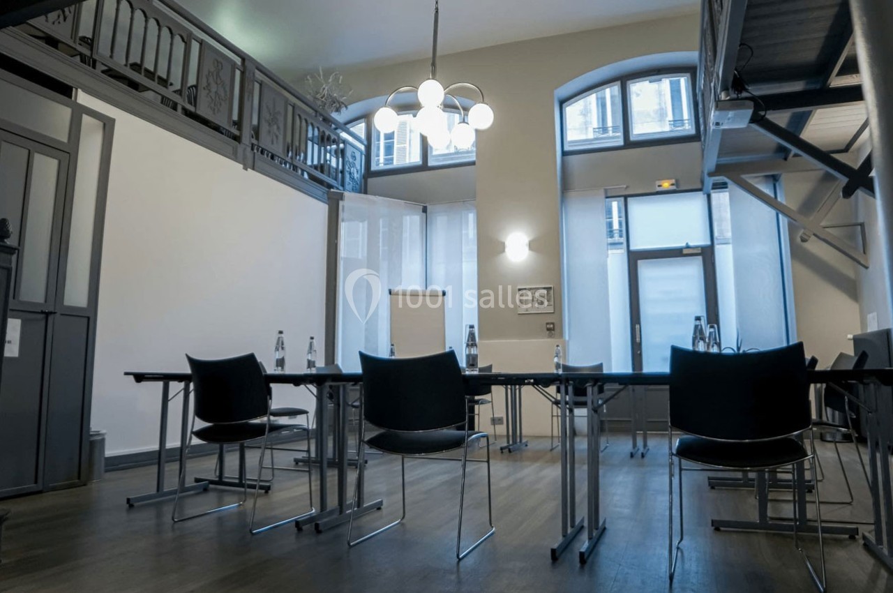 Salle de réunion lumineuse avec grandes fenêtres, tables alignées, chaises noires et bouteilles d'eau disposées.