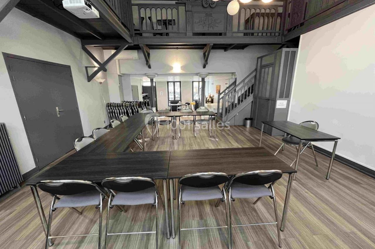 Salle de réunion lumineuse avec tables en U, chaises pliantes et mezzanine visible en arrière-plan.