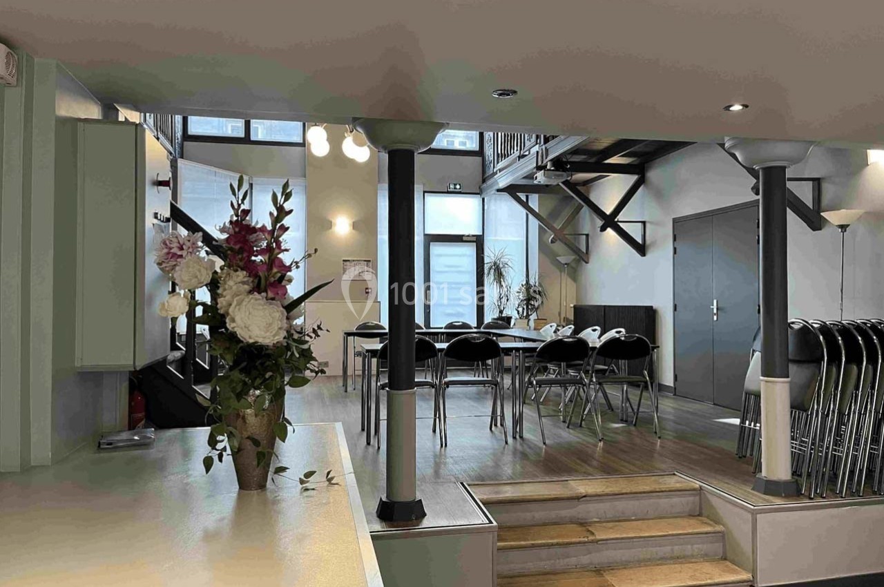 Salle lumineuse avec tables, chaises empilées, escalier métallique et vase de fleurs près d'une colonne.