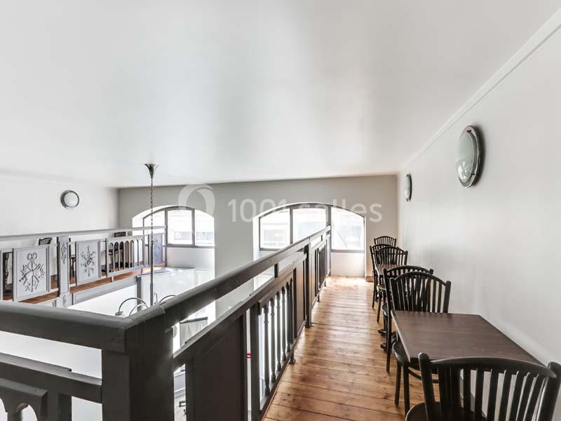 Passerelle en bois avec tables et chaises, surplombant un espace lumineux avec de grandes fenêtres et des murs clairs.