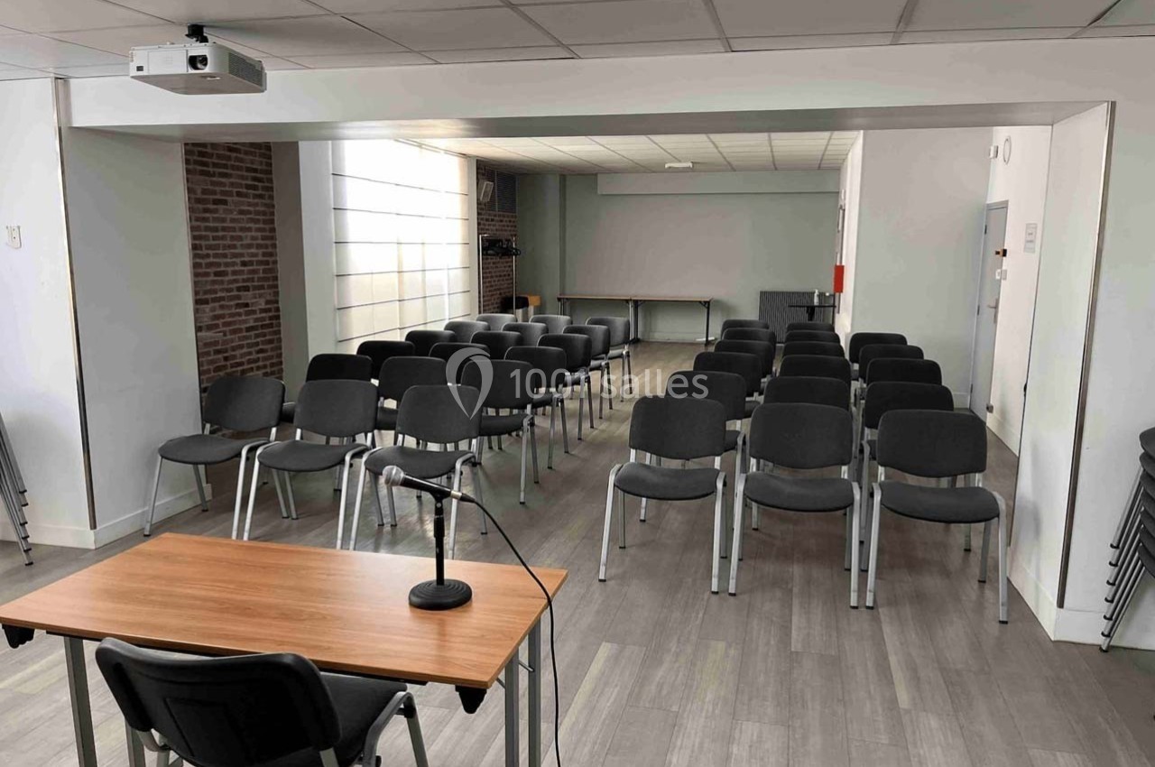 Salle de conférence avec des rangées de chaises grises, une table avec un micro et un écran blanc en fond.