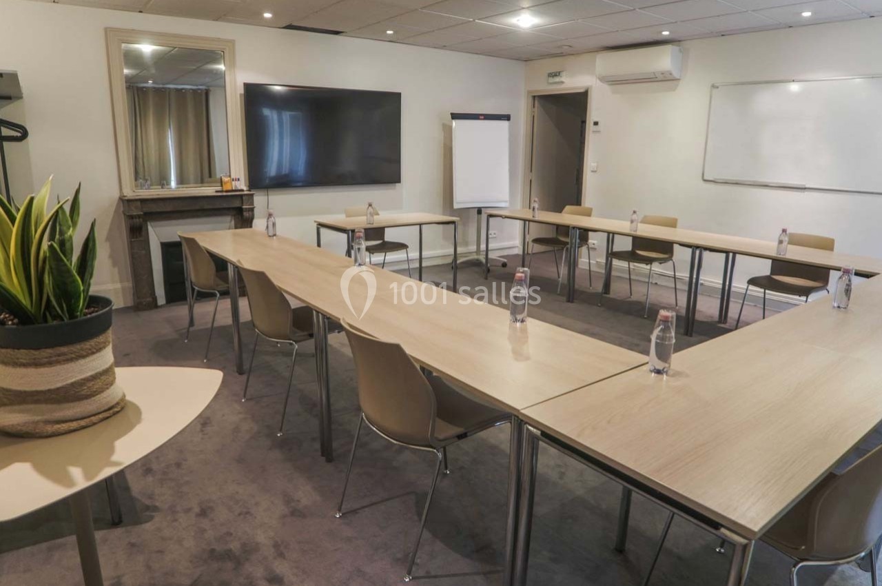 Salle de réunion lumineuse avec tables disposées en U, chaises, tableau blanc, écran et plantes décoratives.