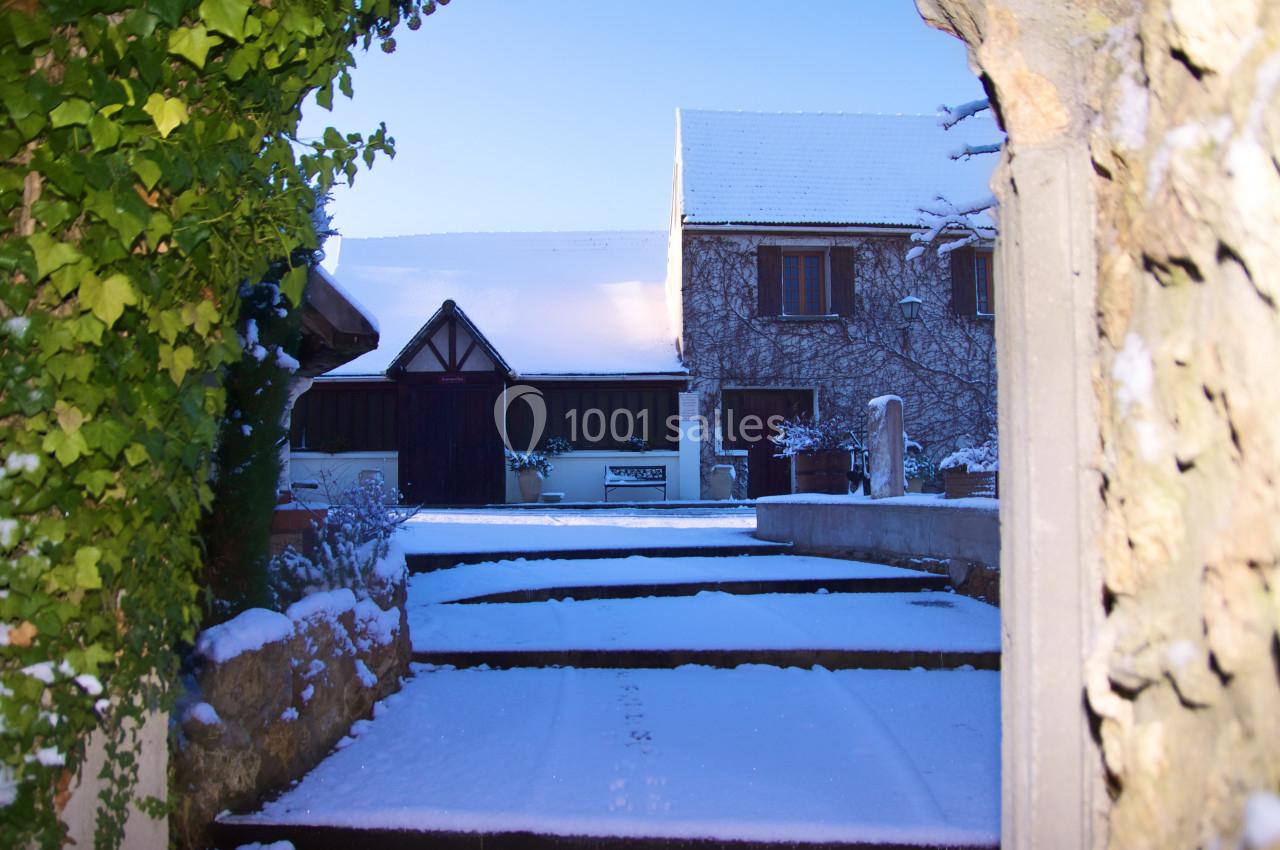 Cour enneigée entourée de murs en pierre, avec une maison en arrière-plan sous un ciel bleu clair.