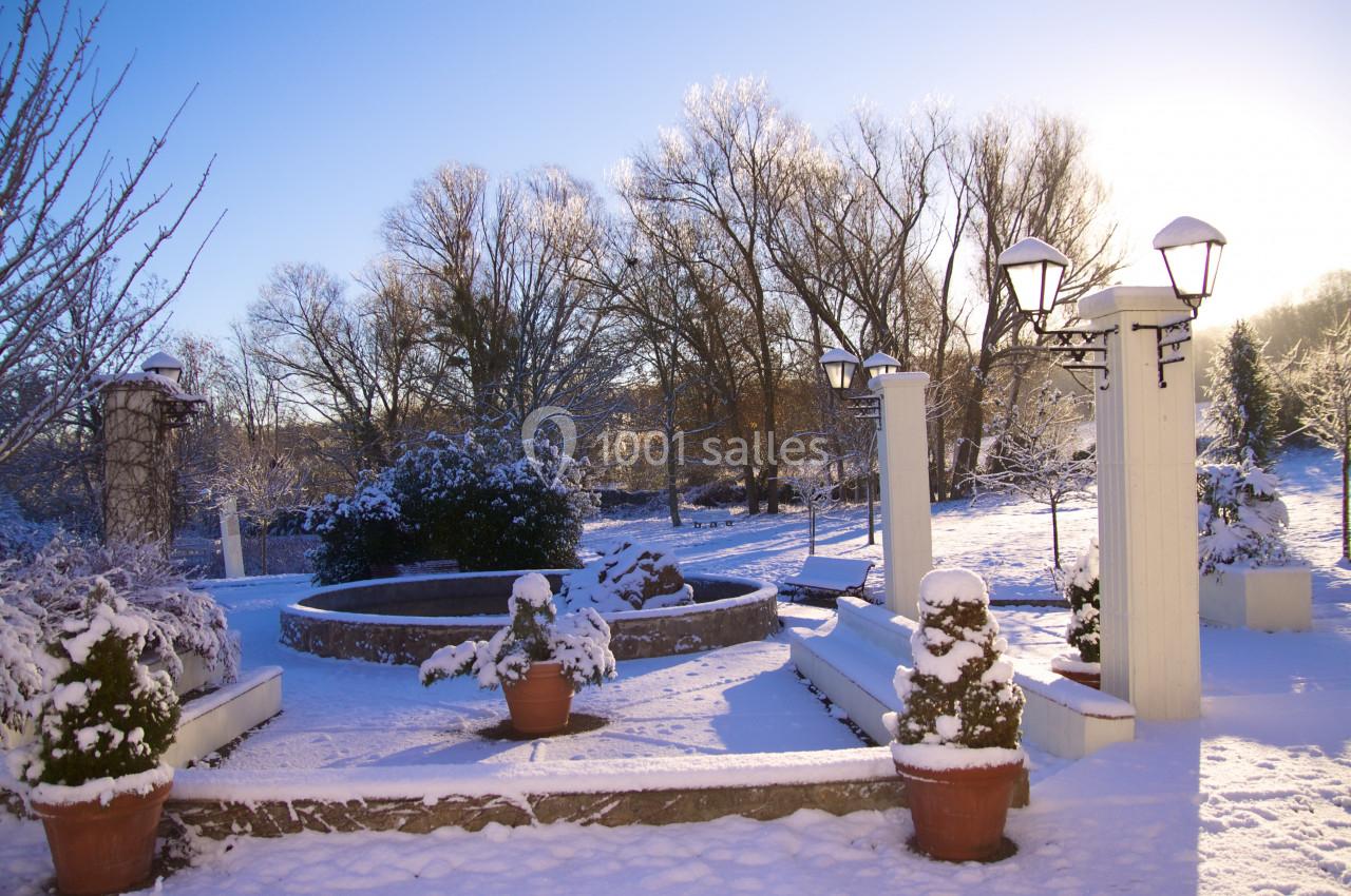Jardin enneigé avec fontaine centrale, arbres dénudés et lampadaires sous un ciel dégagé ensoleillé.