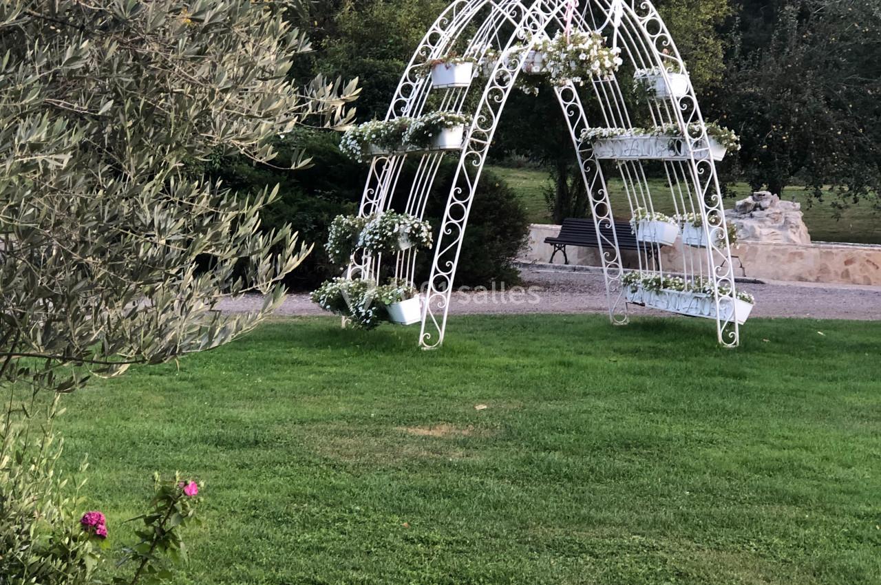 Structure métallique blanche en forme d'arche sur une pelouse verte, entourée d'arbres et de fleurs.