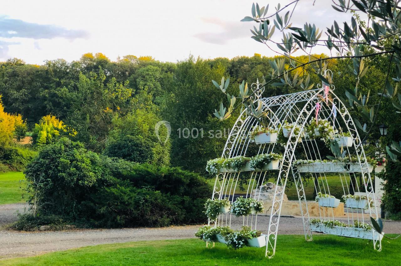 Structure métallique blanche décorée de pots de fleurs, située dans un jardin verdoyant avec des arbres en arrière-plan.