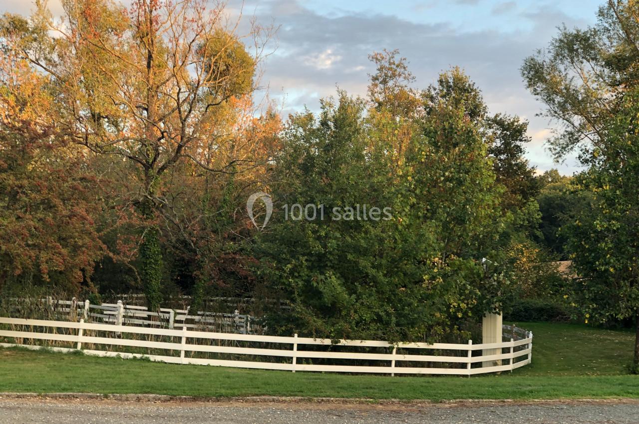 Clôture blanche entourant des arbres et buissons dans un paysage naturel sous un ciel partiellement nuageux.