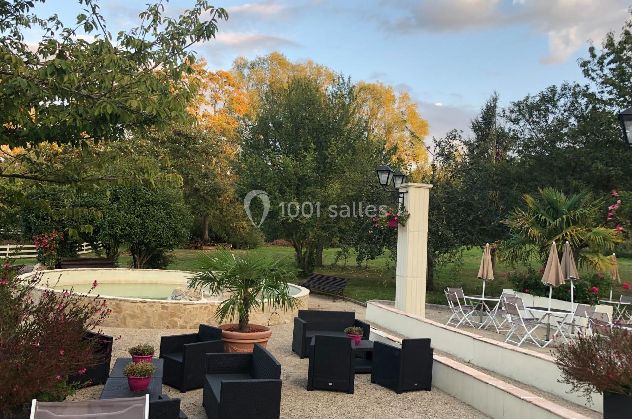 Espace extérieur avec mobilier de jardin, fontaine, végétation luxuriante et ciel partiellement nuageux en arrière-plan.