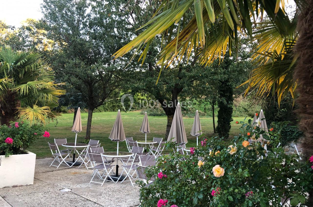 Terrasse extérieure avec tables, chaises pliantes et parasols, entourée de palmiers, rosiers et arbres verdoyants.