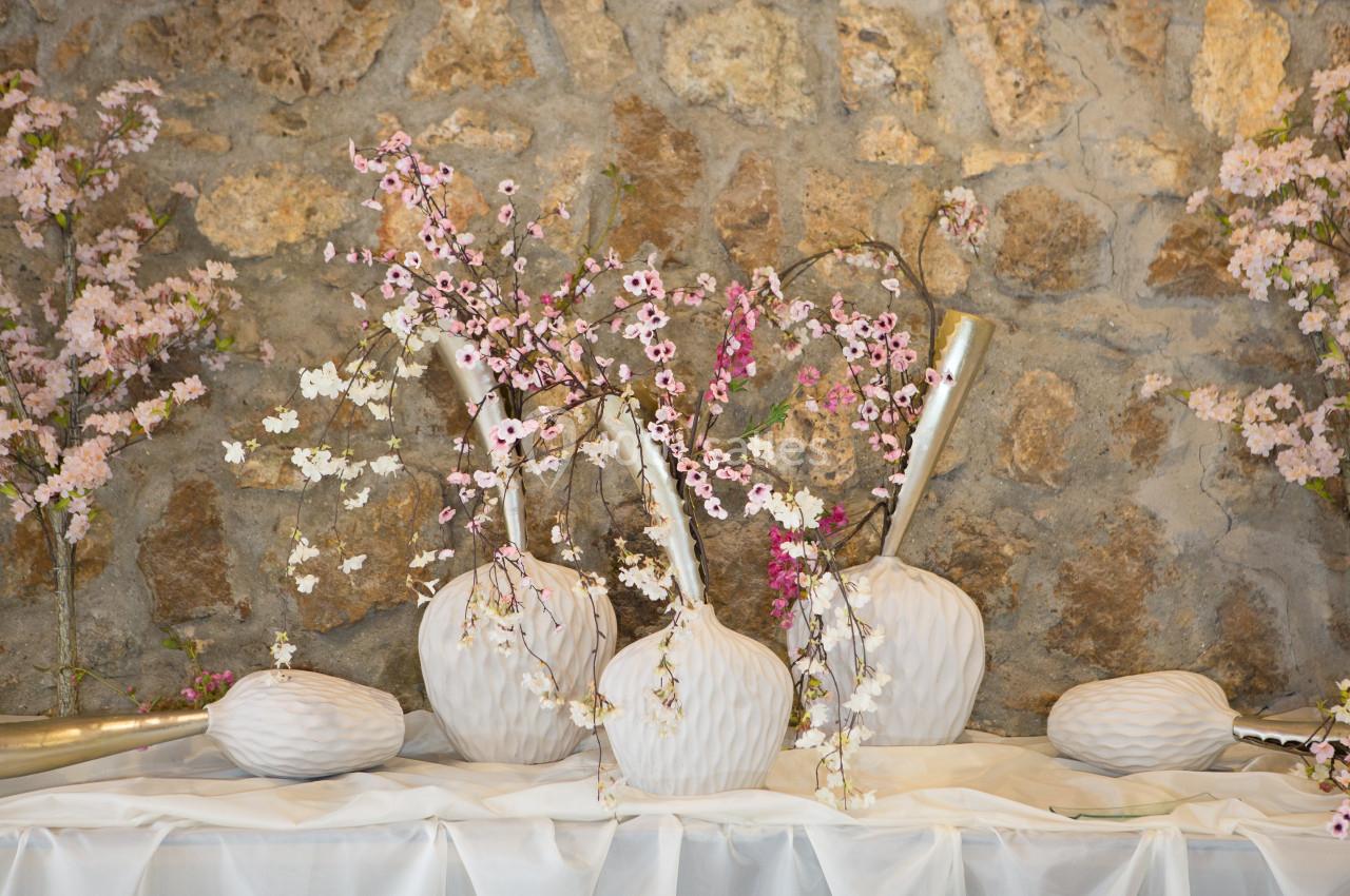 Branches de fleurs de cerisier disposées dans des vases blancs texturés devant un mur en pierre.