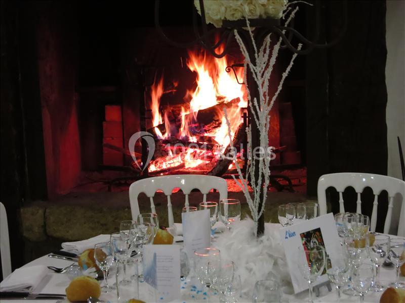 Table dressée avec vaisselle et décorations blanches devant une cheminée allumée diffusant une lumière chaleureuse.