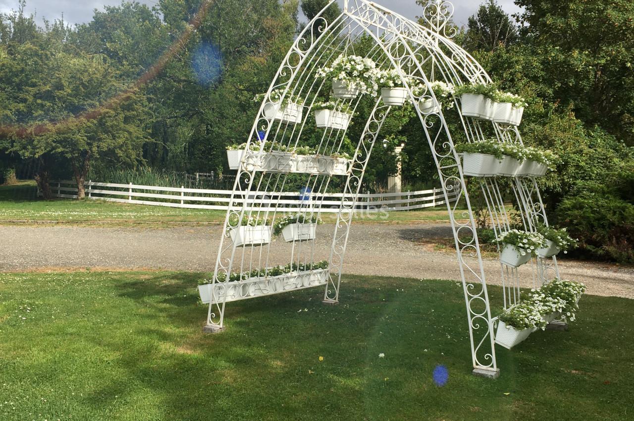 Arche blanche décorative en métal avec des fleurs blanches, installée sur une pelouse dans un jardin verdoyant.