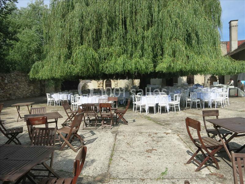 Tables et chaises en bois et en métal disposées sous un grand saule pleureur dans une cour pavée.