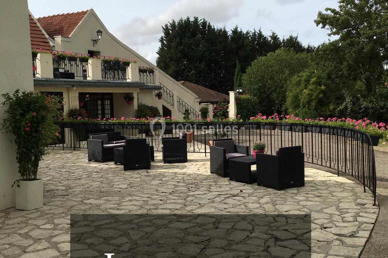 Terrasse en pierre avec fauteuils noirs, entourée de fleurs et d'arbres, devant un bâtiment à balcons fleuris.
