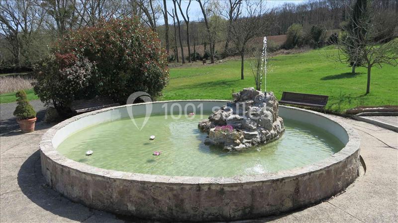 Bassin circulaire avec une fontaine centrale en pierre, entouré de bancs et d'un espace vert arboré.