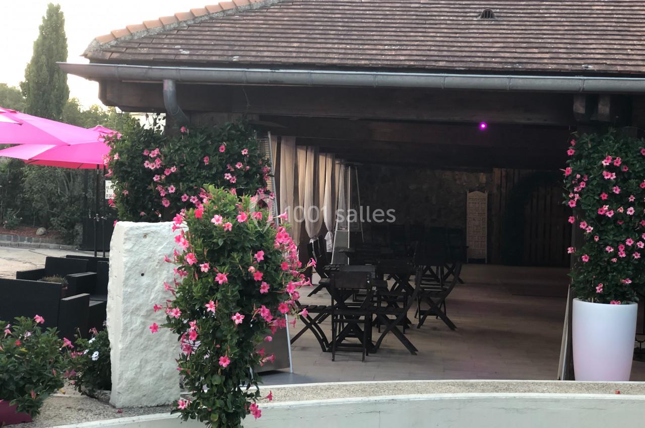 Terrasse couverte avec tables et chaises en bois, entourée de plantes fleuries et éclairée par une lumière violette.
