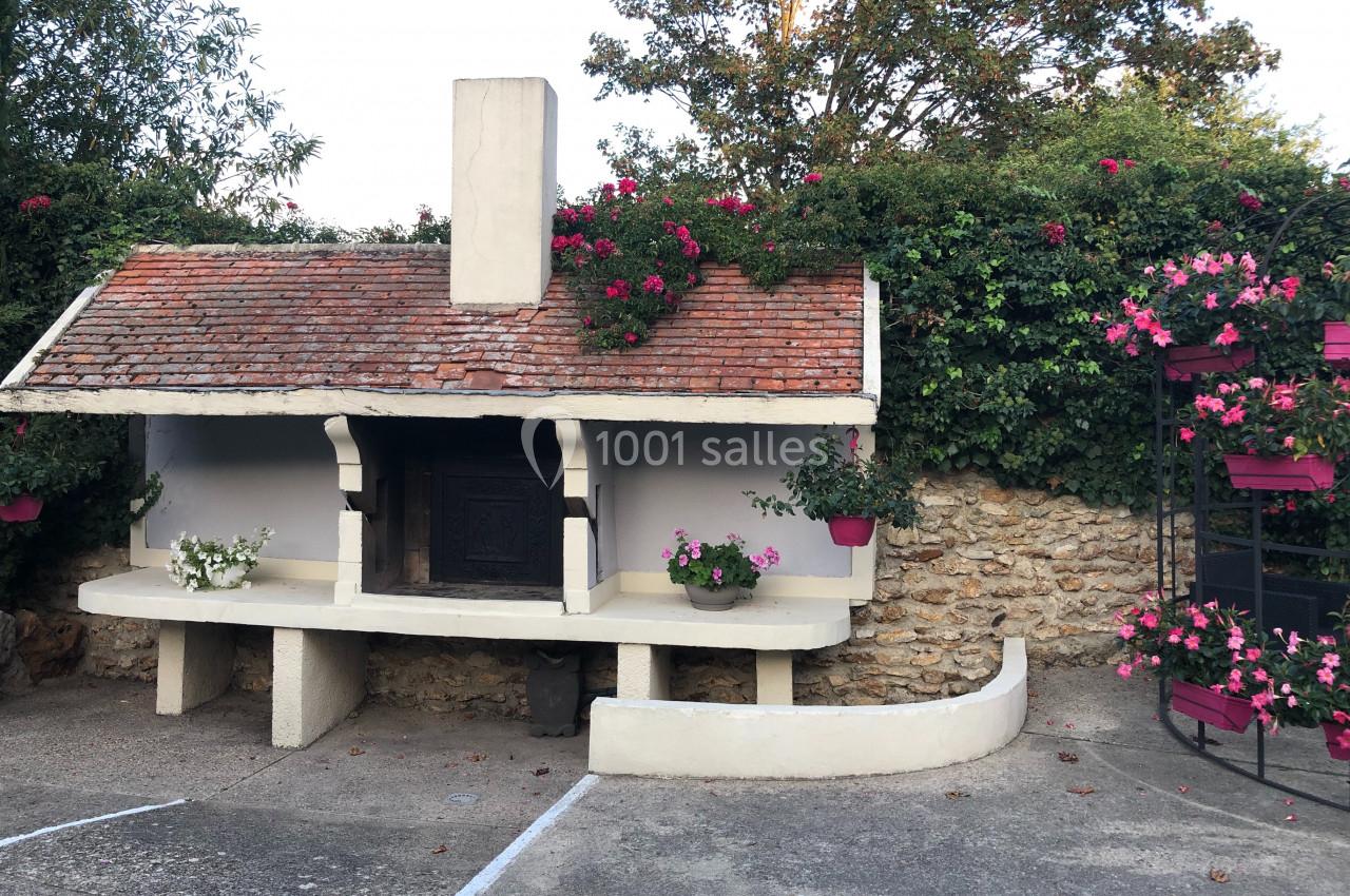 Barbecue en pierre avec toit en tuiles, entouré de fleurs et de verdure, dans une cour extérieure pavée.