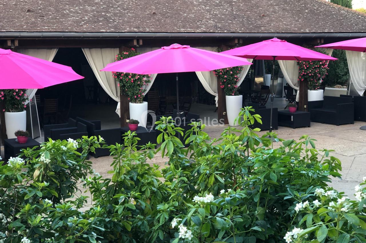 Terrasse avec parasols roses, mobilier de jardin noir, pots de fleurs blancs et végétation en premier plan.