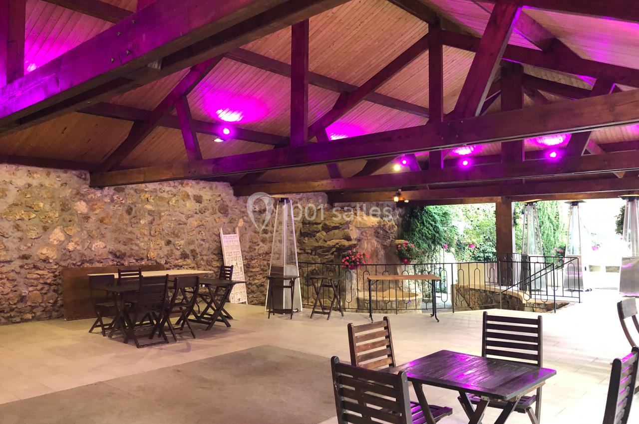 Salle avec charpente en bois, murs en pierre, éclairage violet et mobilier en bois disposé sur un sol clair.